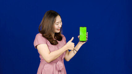 beautiful asian woman pointing green screen in handphone isolated blue background