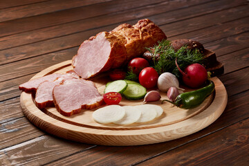 Ham garnished with vegetables, fruits and herbs with sauces and bread on a wooden plate on a dark wooden table.