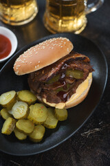 Black plate with brisket sandwich, pickles and beer mugs in the background, vertical shot, close-up