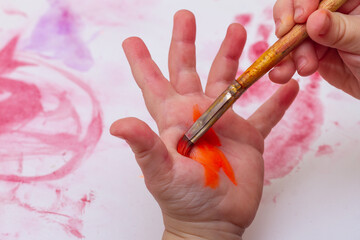 The child paints his hand with a brush in orange.