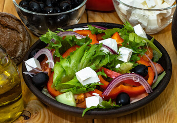 Greek salad in a dark ceramic plate. The plate is surrounded by ingredients: feta cheese, black olives, olive oil. Next to it is a dark ceramic cup and a loaf of bread.