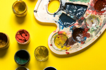 dirty palette and open jars of gouache paints on a yellow background. place for text. children's...