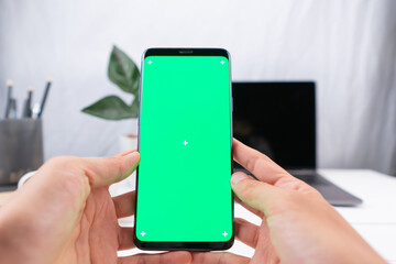 Close up hand holding a smartphone with Mockup, a blank green screen. with laptop and pencil in the white coffee shop