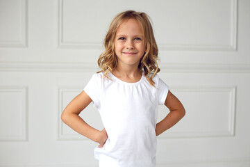 Front portrait of a little girl smiling and looking at the camera, dressed in a white T-shirt, holding her hands on her waist, on a white background.