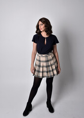 Full length portrait of pretty brunette woman wearing tartan skirt and blouse.  Standing pose on the ground,  against a  studio background.