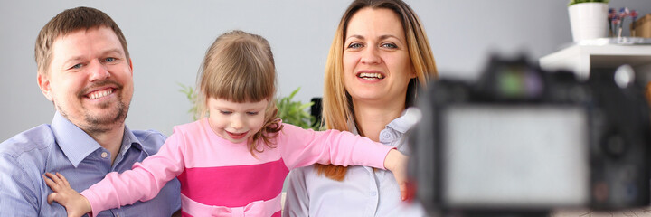 Waist up portrait of smiling parents and little girl sitting at couch and using camera and tripod for taking picture