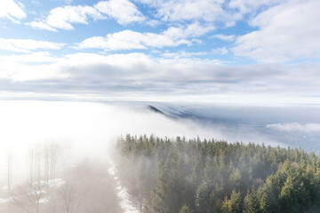 Obraz premium The tops of the hills during a sunny afternoon covered with thick fog which passes between the treetops in the background of blue sky.
