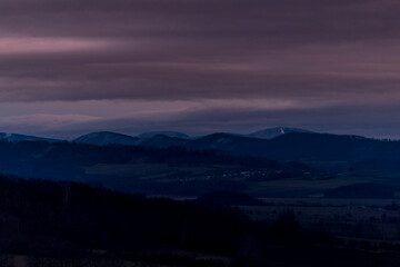 Sunset dark sky with clouds over the mountainous landscape of Pustevny lying under the fog in a dark color.