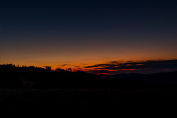 Field where grass grows overlooking the surrounding hills and treetops during sunset on an orange horizon overlooking the landscape and nature.