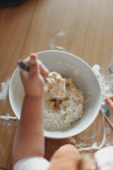 hands of children stir a dough in the white bowl with spoon, on the wooden table in the kitchen. The process of mixing ingredients for pastry. Closeup, selective focus.