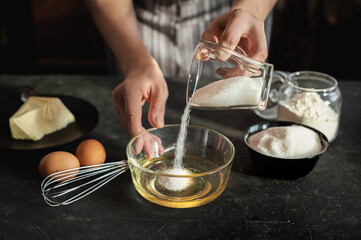Process cooking dough. Ingredients for the dough on a dark background. The woman pours the sugar from cup to bowl with egg mixture.
