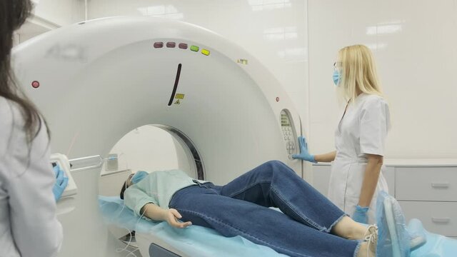 Female patient is undergoing CT or MRI scan under supervision of two qualified radiologists in modern medical clinic. Patient lying on a CT scan bed, in protective mask moving inside the machine.