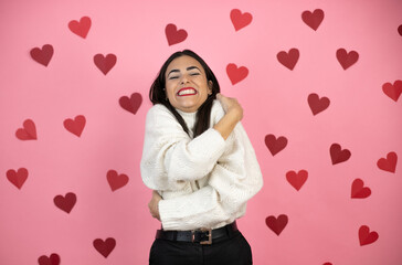 Young beautiful woman over pink background with harts hugging oneself happy and positive, smiling confident