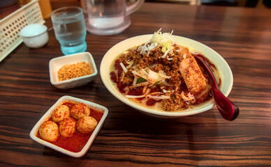 A bowl of tantanmen noodles with sides of fried garlic bits and spicy marinated boiled quail eggs on a wooden table in a restaurant