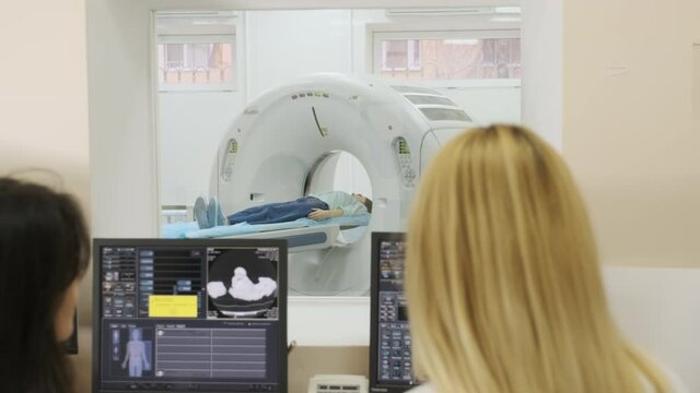 Two qualified radiologists examines female patient who is lying under CT or MRI scanner. Computer screen with result diagnosis image. View through glass window in a modern clinic.