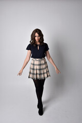 Full length portrait of pretty brunette woman wearing tartan skirt and blouse.  Standing pose on the ground,  against a  studio background.