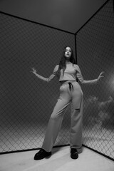 Young woman with long straight brunette hair in sporty clothes standing against grid fence and pink background. Full length fashion portrait