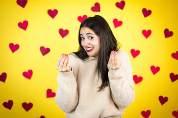 Young caucasian woman over yellow background with red hearts doing money gesture with hands, asking for salary payment, millionaire business