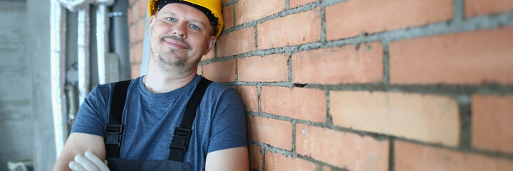 Waist up portrait of smiling bearded man in yellow hard hat standing and leaning against brick wall