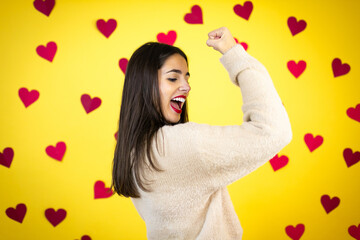 Young caucasian woman over yellow background with red hearts feeling happy, satisfied and powerful, flexing fit and muscular biceps, looking strong after the gym