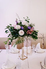 Table covered with a tablecloth and served with dishes and cutlery. Festive table decorated with composition of flowers and greenery in the wedding banquet hall.