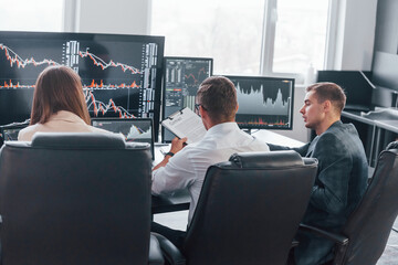Holds document in hands. Team of stockbrokers works in modern office with many display screens