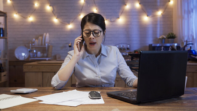 Angry Female Manager Yelling At Mobile Phone At Dining Table In Late Night Home Kitchen. Girl Worker Disagree And Argue With Team Partner On Cellphone While Working On Deadline Project In Evening.