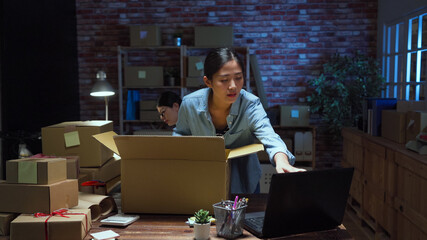 Startup small business entrepreneur SME. asian woman packing cloth in cardboard box and checking customer order online on laptop computer. lady coworker sitting in background overtime working.