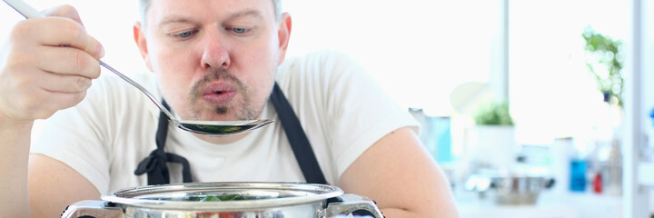 Male in apron is tasting soup from pot with ladle while preparing dinner at home