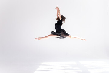 A slender girl in a black sports swimsuit does a split in a jump on a white isolated background. Girl athlete doing splits in the air on a white isolated background with place for text or logo.