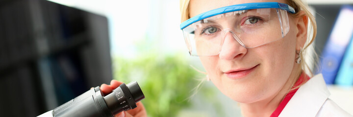 Cheerful female person keeping smile on her face while standing near microscope and looking at camera
