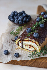 Sponge cake roll with chocolate and cream cheese decorated with chocolate glaze, blueberry and rosemary on parchment, light background. Biscuit swiss roll.