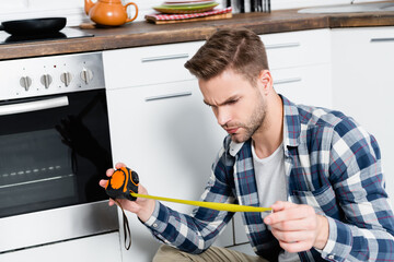 serious young man looking at tape measure near oven in kitchen