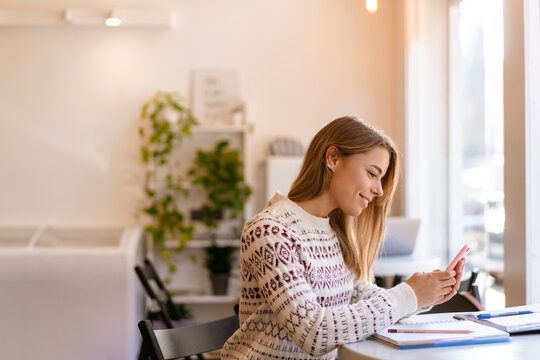 Smiling student girl using mobile phone while doing homework in cafe