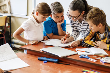 Female teacher helps school kids to finish they lesson.They sitting all together at one desk.