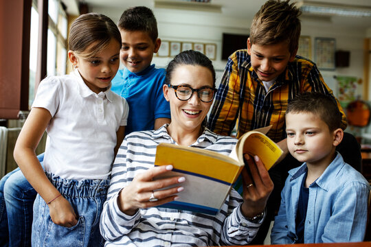 Female Teacher Reading A Book To Small Group Of School Kids In Classroom.	
