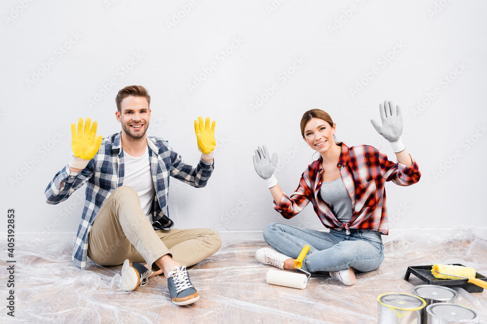 Wall mural full length of happy young couple looking at camera and showing gloves while sitting on floor at hom