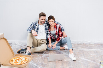 full length of happy young couple hugging and watching film on laptop while sitting on floor near box with pizza at home