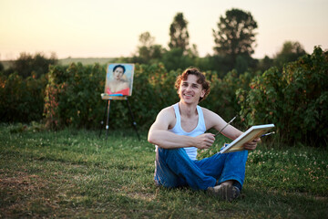 Young male artist, wearing torn jeans and white t-shirt, sitting on ground on green field during sunset, holding empty canvas, thinking. Painting workshop in countryside. Artistic education concept.