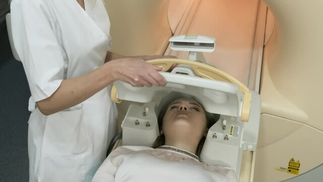 Doctor radiologist prepares female patient for brain MRI scanning, puts modern equipment, coil on the patient's head.