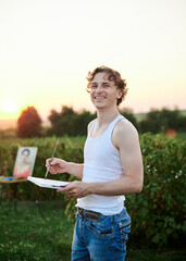 Young male artist, wearing torn jeans and white t-shirt, standing on green field during sunset, holding empty canvas, thinking. Painting workshop in countryside. Artistic education concept.