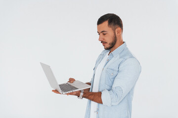 Uses laptop. Young handsome man standing indoors against white background