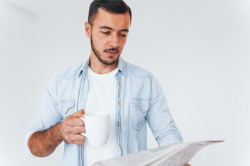 Reads newspaper. Young handsome man standing indoors against white background