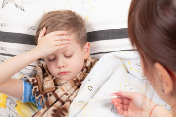 a little boy a child wrapped in a scarf is ill at home, lies in bed and holds on to his forehead, mom gives him a thermometer