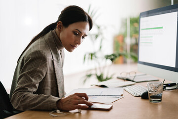 Serious woman using cellphone while working with computer and documents