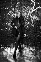 Sexy brunette woman with long wavy hair in black blazer, net body, short leather shorts, tights and shoes standing and posing against shining silver background with stars. Full length portrait