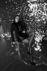 Elegant sexy woman in black net body, blazer, tights sitting on glass chair in festive interior with shining silver wall and party lightning. Fashion portrait
