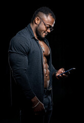 Strong athlete in dark jeans poses to the camera on black background. Unbuttoned black shirt and perfect abs. Portrait of an afro american bodybuilder with phone in hands.
