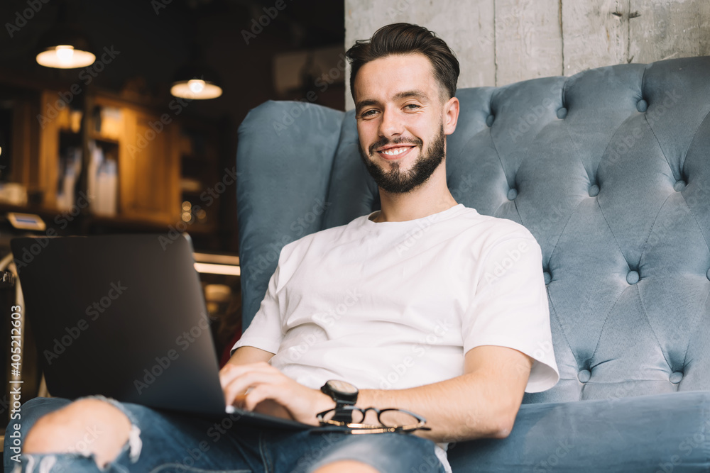 Poster portrait of skilled hipster guy smiling at camera while doing laptop distance job in coworking space