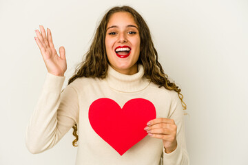 Young caucasian woman holding a heart valentines day shape isolated receiving a pleasant surprise, excited and raising hands.